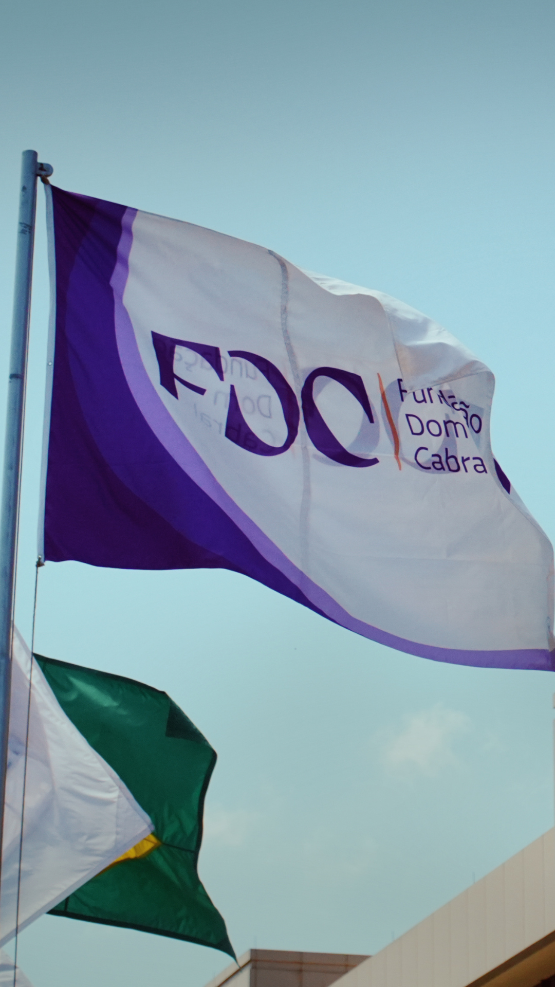 FDC (Fundação Dom Cabral) flag flying in front of a modern campus building, with the Brazilian flag and a clear blue sky in the background.