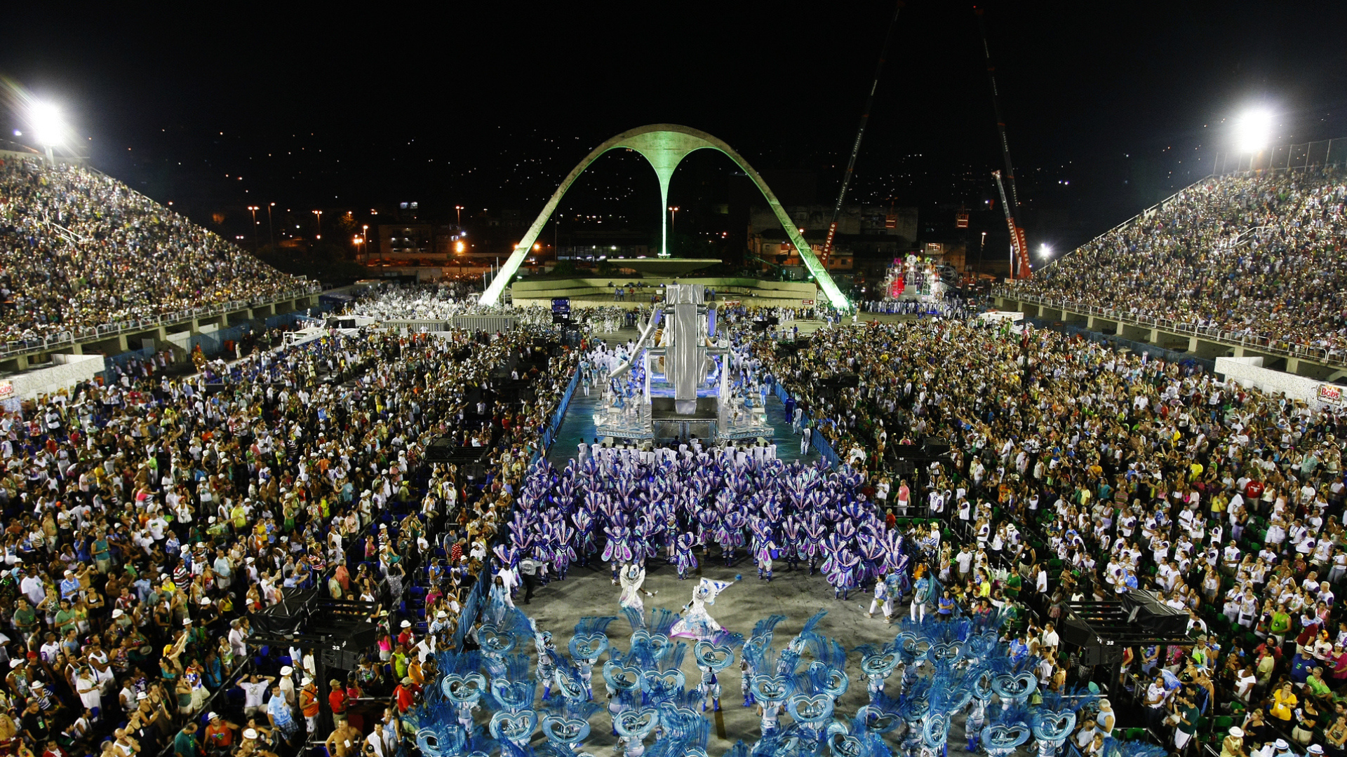 Nighttime view of a large outdoor event with thousands of people gathered around a central performance area beneath a prominent arch-shaped monument. Bright stadium lights illuminate the crowd, with city lights visible in the background.