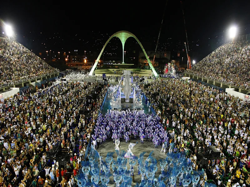Nighttime view of a large outdoor event with thousands of people gathered around a central performance area beneath a prominent arch-shaped monument. Bright stadium lights illuminate the crowd, with city lights visible in the background.
