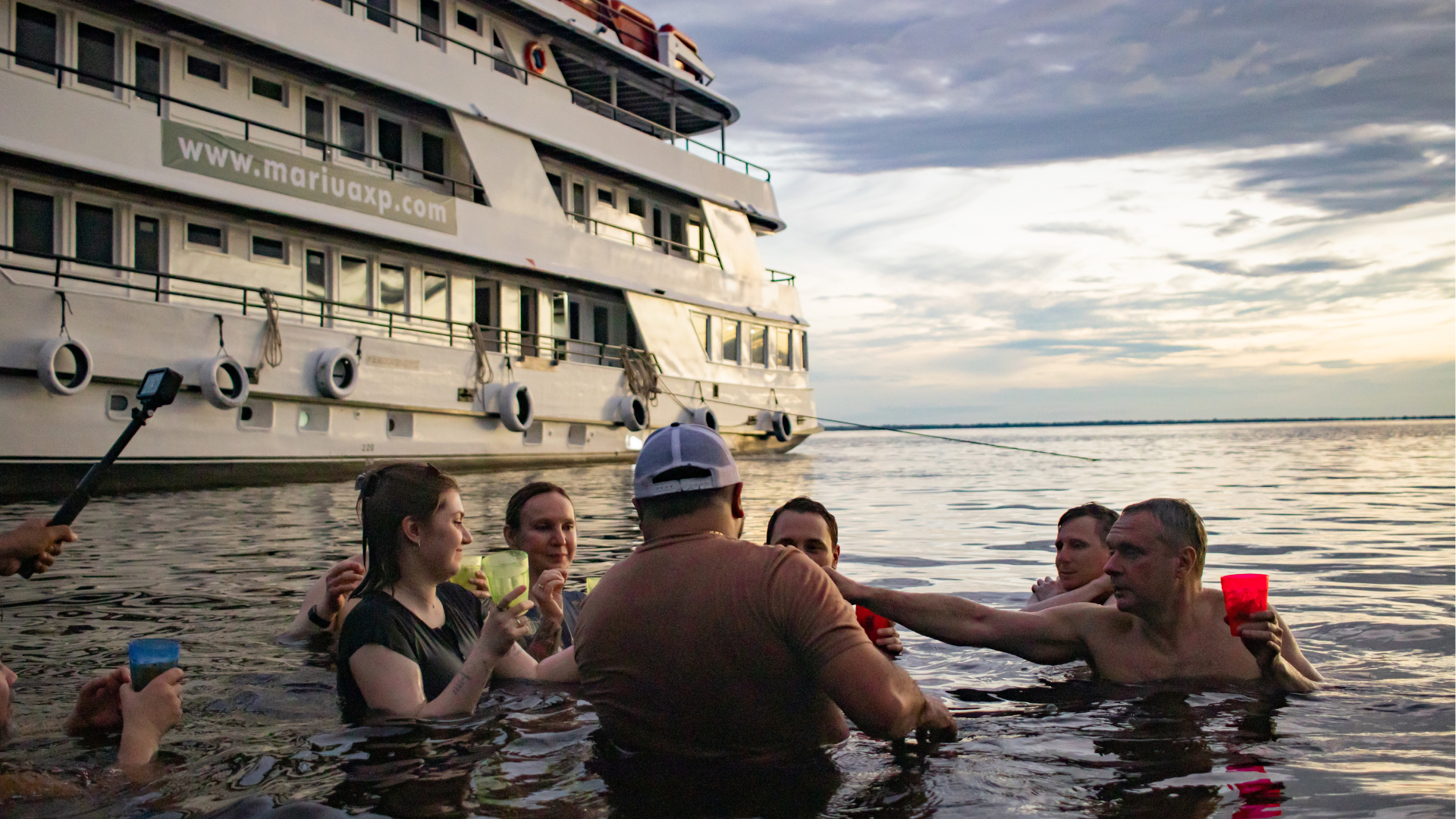People gathered in a river at sunset beside a large riverboat, socializing and sharing drinks while partially submerged in the water.