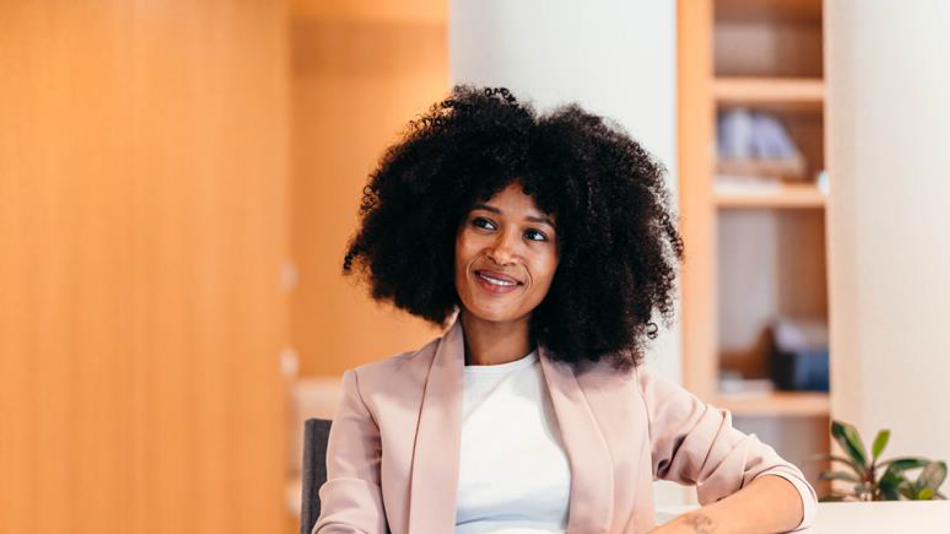 Mulher negra com cabelos cacheados, usando blazer rosa claro e camiseta branca, sorrindo em ambiente de escritório moderno, promovendo empoderamento feminino.