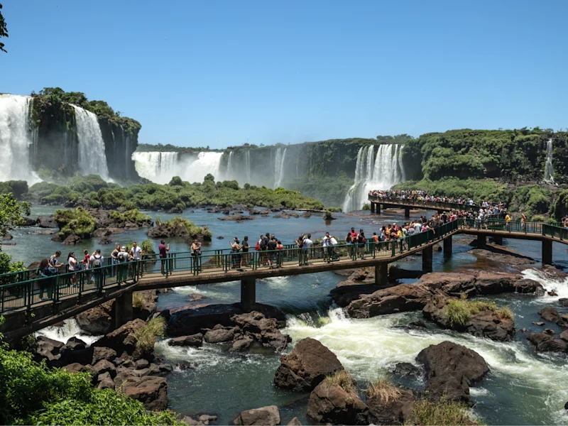 Wide view of a waterfall system cascading into a rocky river, with visitors walking along elevated boardwalks and viewing platforms surrounded by lush green vegetation under a clear blue sky.