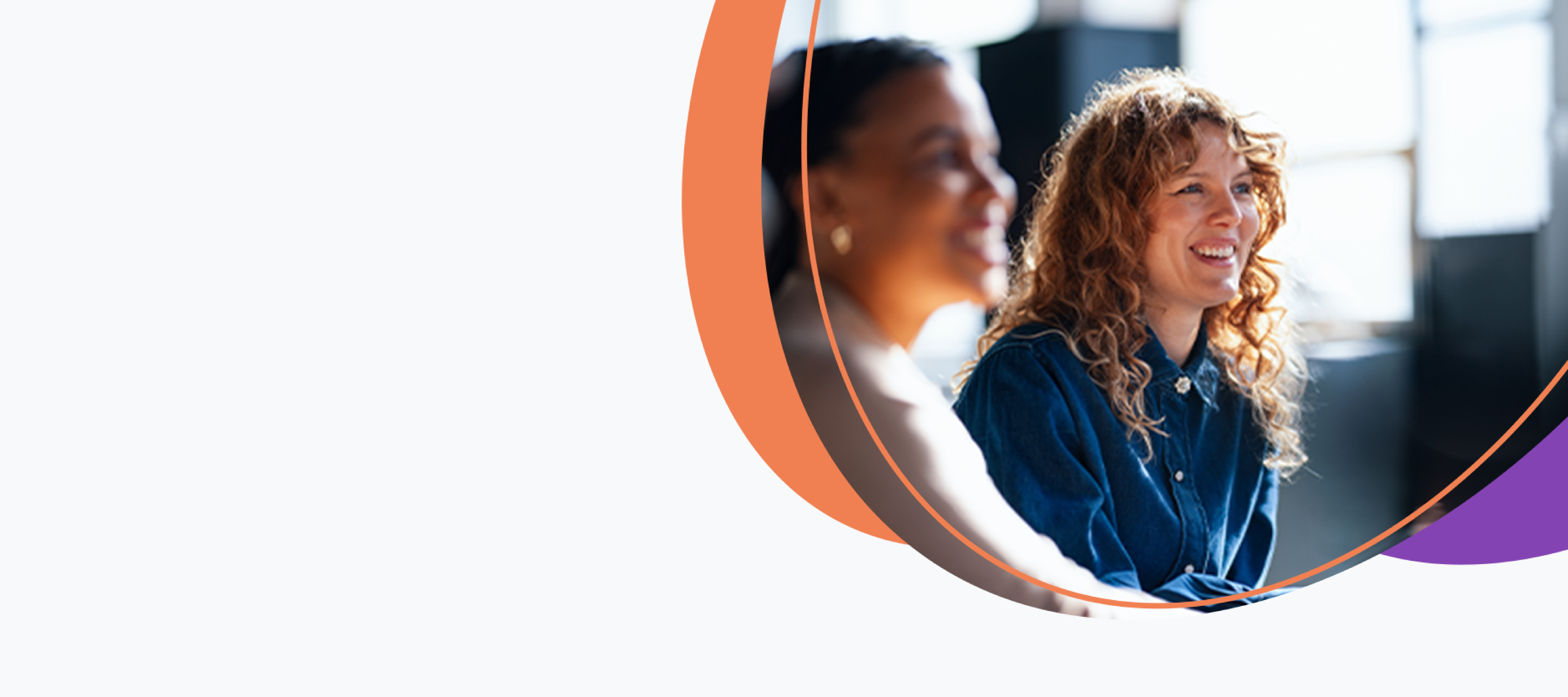 Duas mulheres com roupa social, sorrindo, uma em primeiro plano embaçado e outra ao fundo, em ambiente desfocado corporativo.