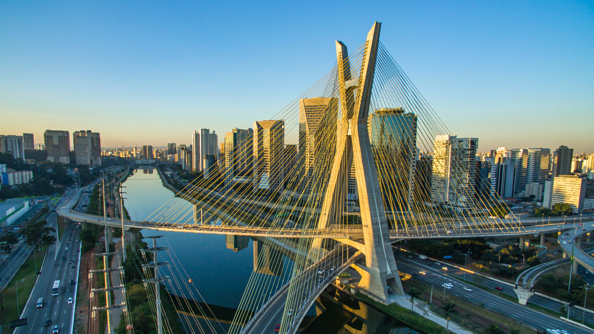 Aerial view of a cable-stayed bridge crossing a river through a modern city skyline at sunset. Distinctive X-shaped towers and radiating cables frame high-rise buildings, highways, and waterways under a clear blue sky.