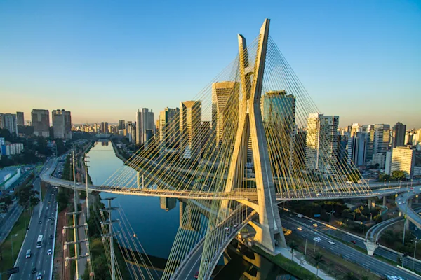 Aerial view of a cable-stayed bridge crossing a river through a modern city skyline at sunset. Distinctive X-shaped towers and radiating cables frame high-rise buildings, highways, and waterways under a clear blue sky.