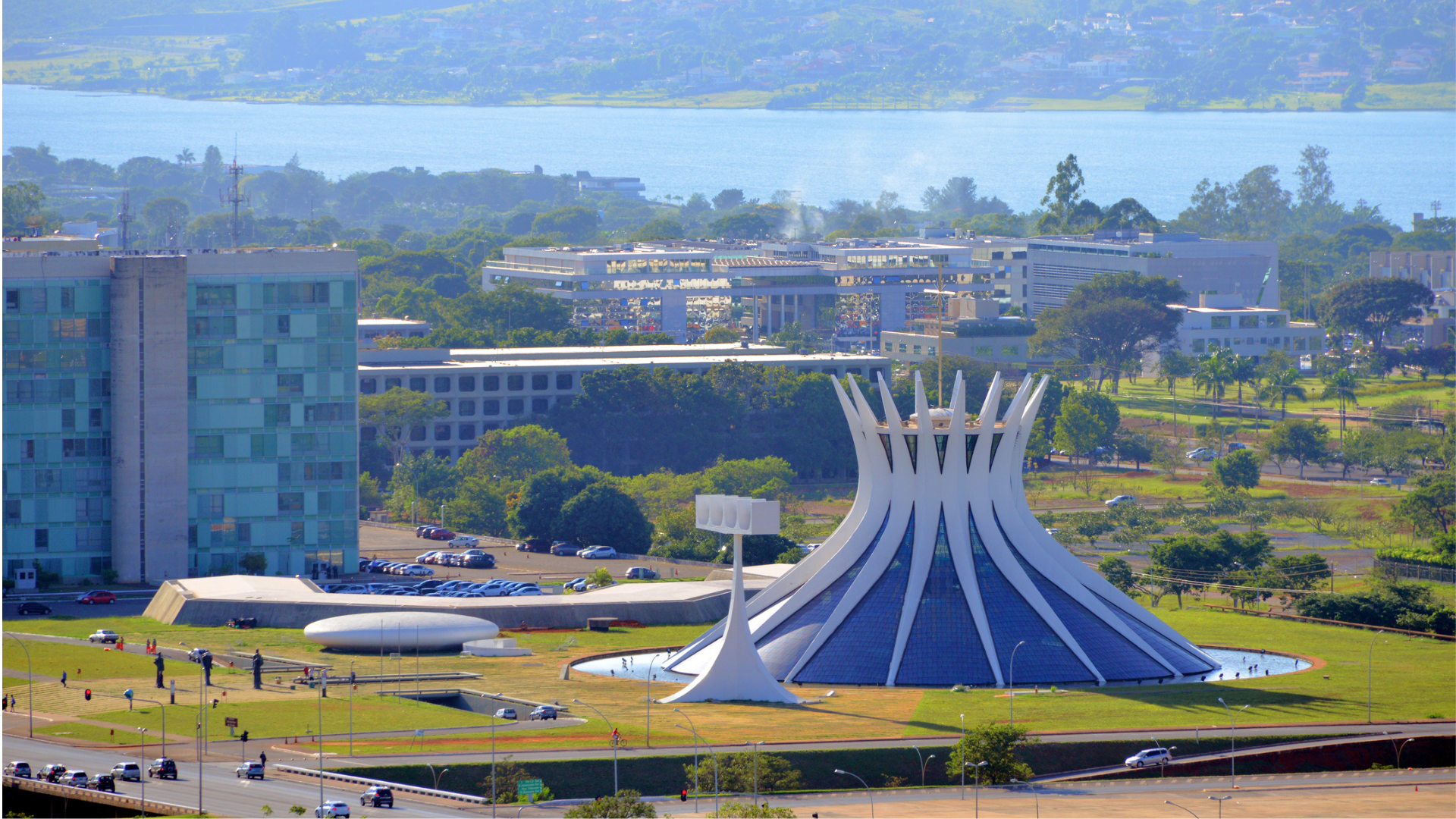 Aerial view of Brasília’s Metropolitan Cathedral surrounded by green lawns and modern government buildings. The iconic white structure stands out against the cityscape, with a lake and distant hills visible in the background.