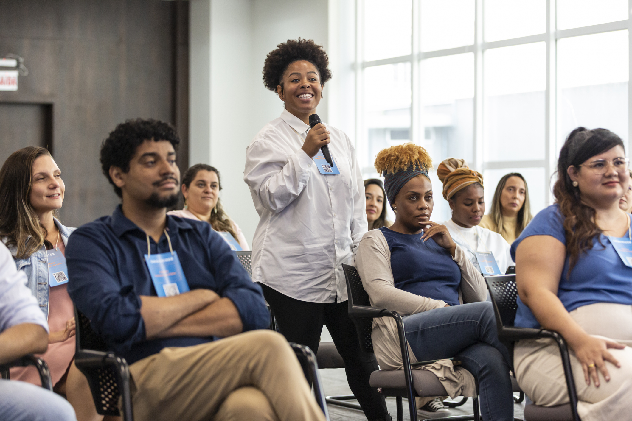 Mulher negra sorridente participando de um painel em uma conferência, com outras pessoas ao redor ouvindo atentamente, evento de diversidade e inclusão.