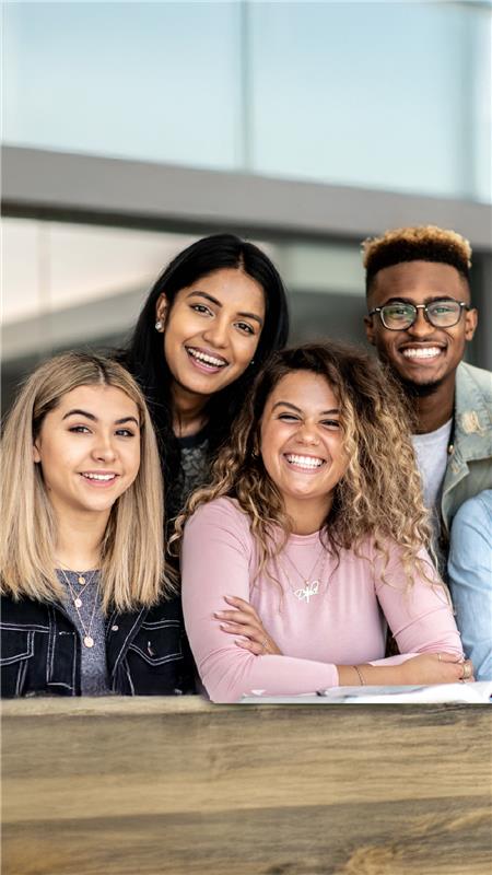 Um grupo de cinco jovens adultos, três mulheres e dois homens, sorrindo e inclinando-se em direção à câmera. Eles transmitem uma sensação de união e camaradagem alegre.