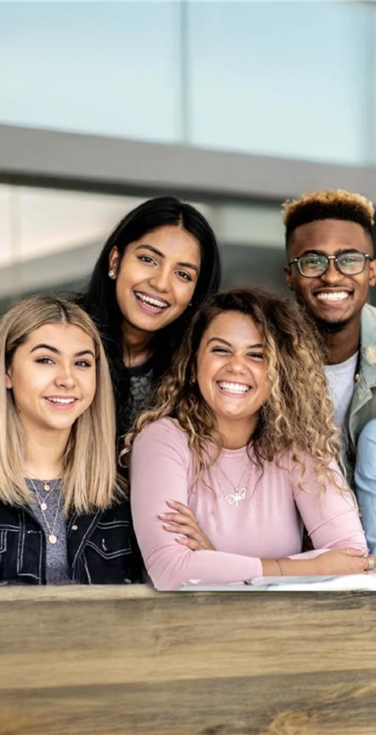 Um grupo de cinco jovens adultos, três mulheres e dois homens, sorrindo e inclinando-se em direção à câmera. Eles transmitem uma sensação de união e camaradagem alegre.