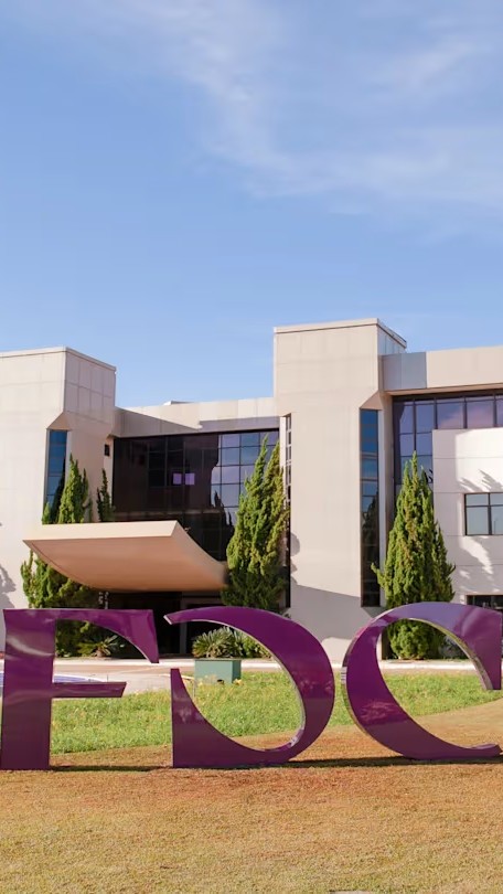 Front view of a modern educational campus building with large windows, landscaped grounds, and a prominent purple FDC sculpture on the lawn under a clear blue sky.