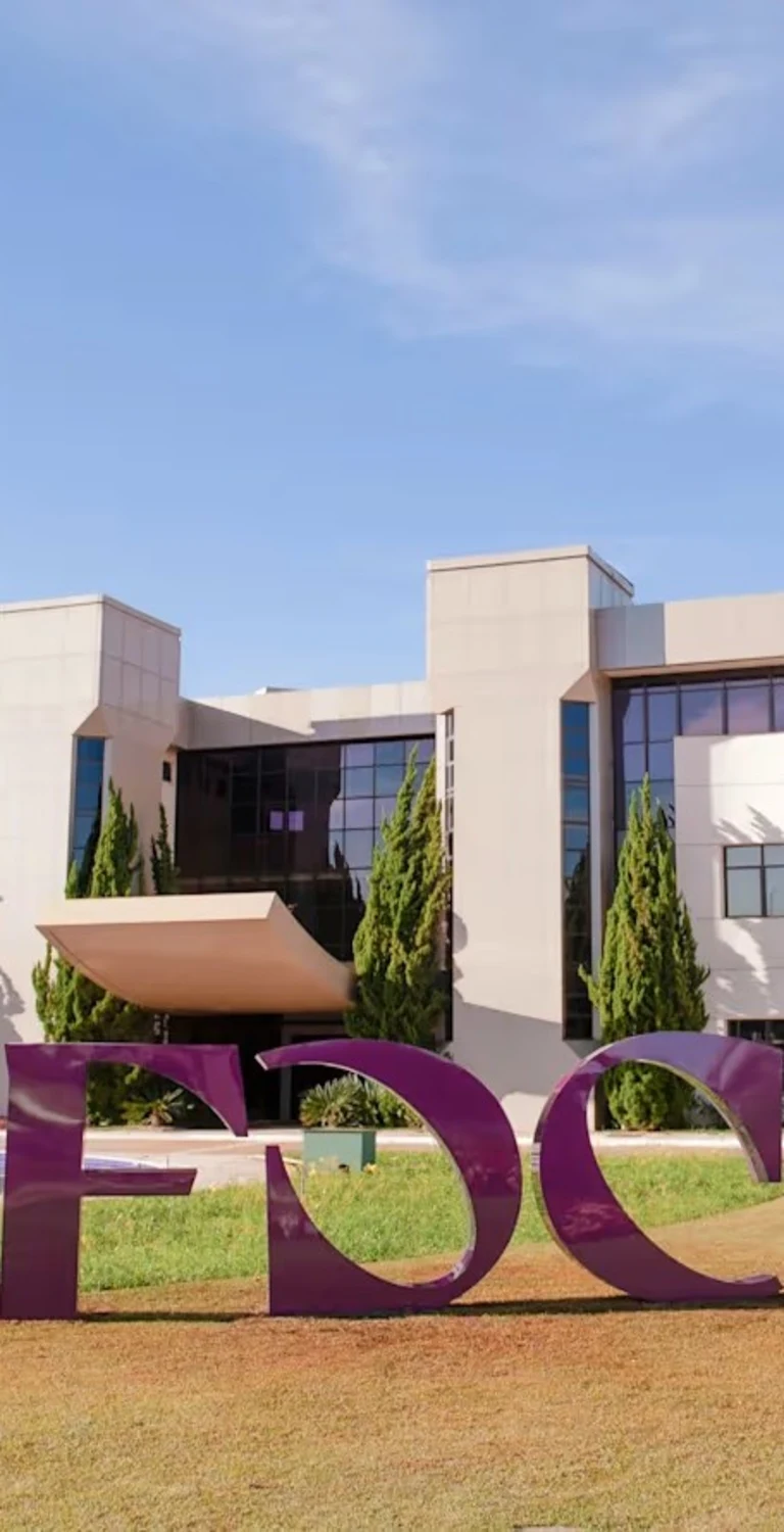 Front view of a modern educational campus building with large windows, landscaped grounds, and a prominent purple FDC sculpture on the lawn under a clear blue sky.