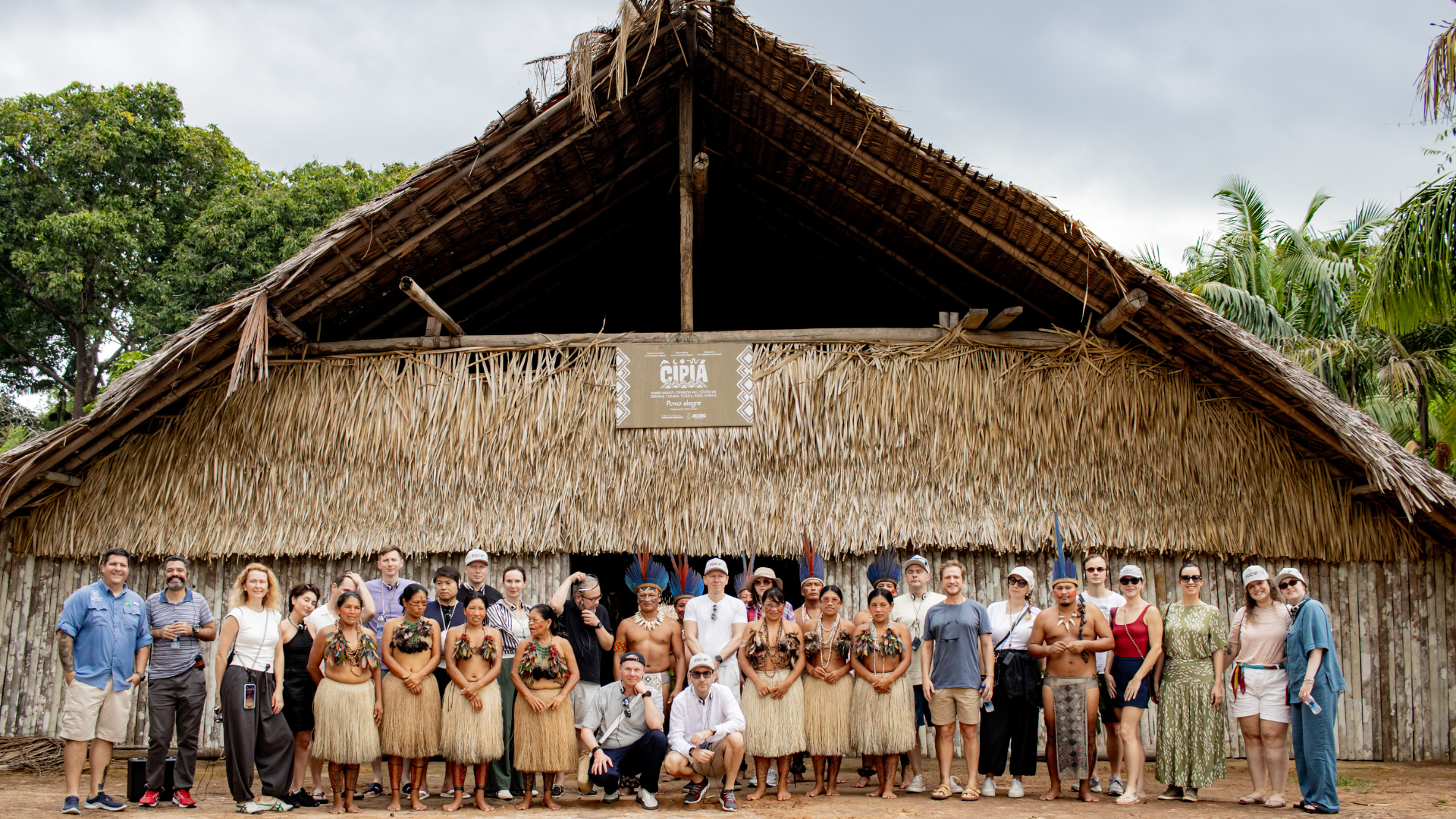 Group photo of visitors and Indigenous community members standing in front of a large traditional thatched maloca, celebrating a cultural gathering in the Amazon rainforest.