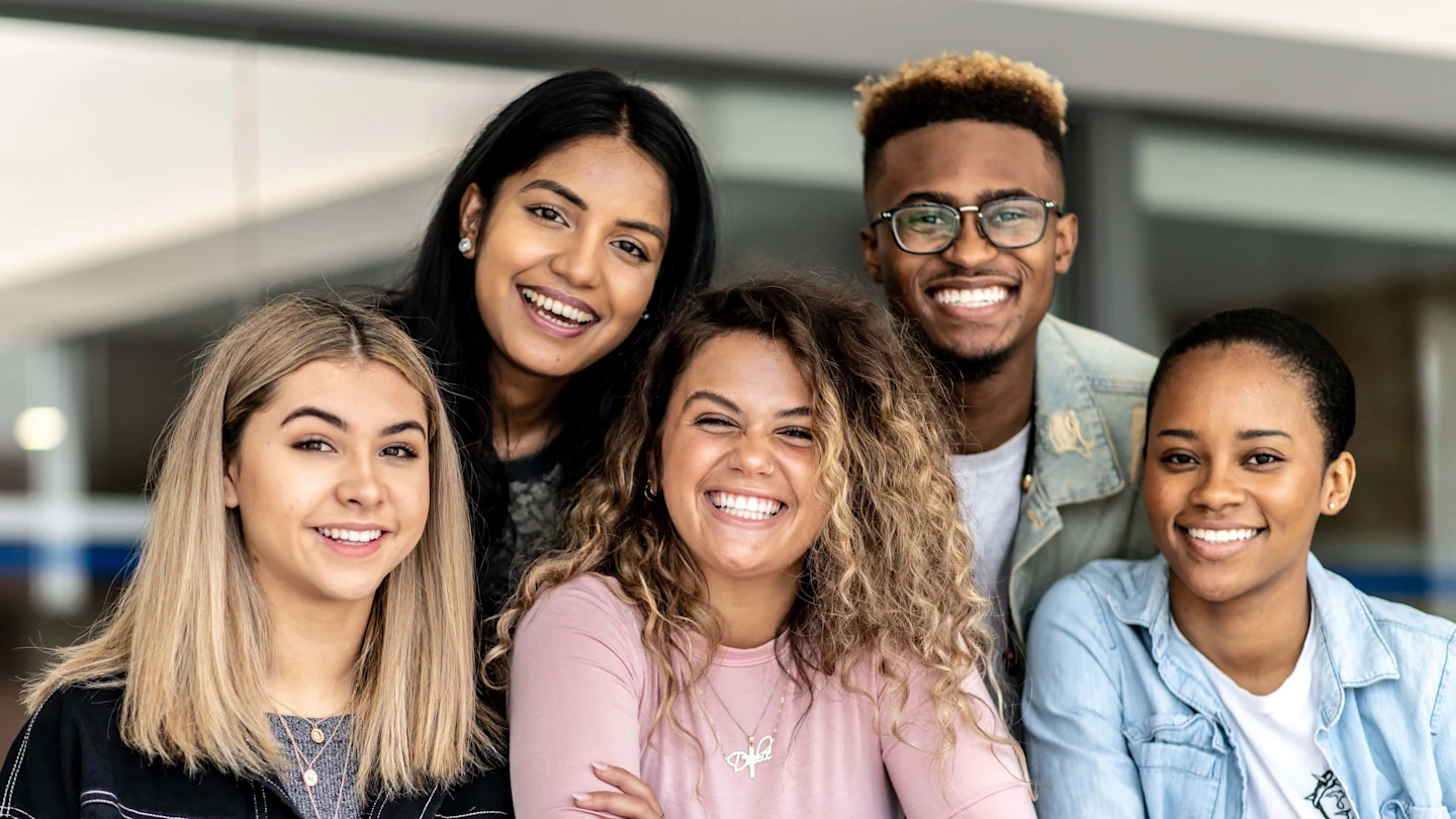 Um grupo de cinco jovens adultos, três mulheres e dois homens, sorrindo e inclinando-se em direção à câmera. Eles transmitem uma sensação de união e camaradagem alegre.