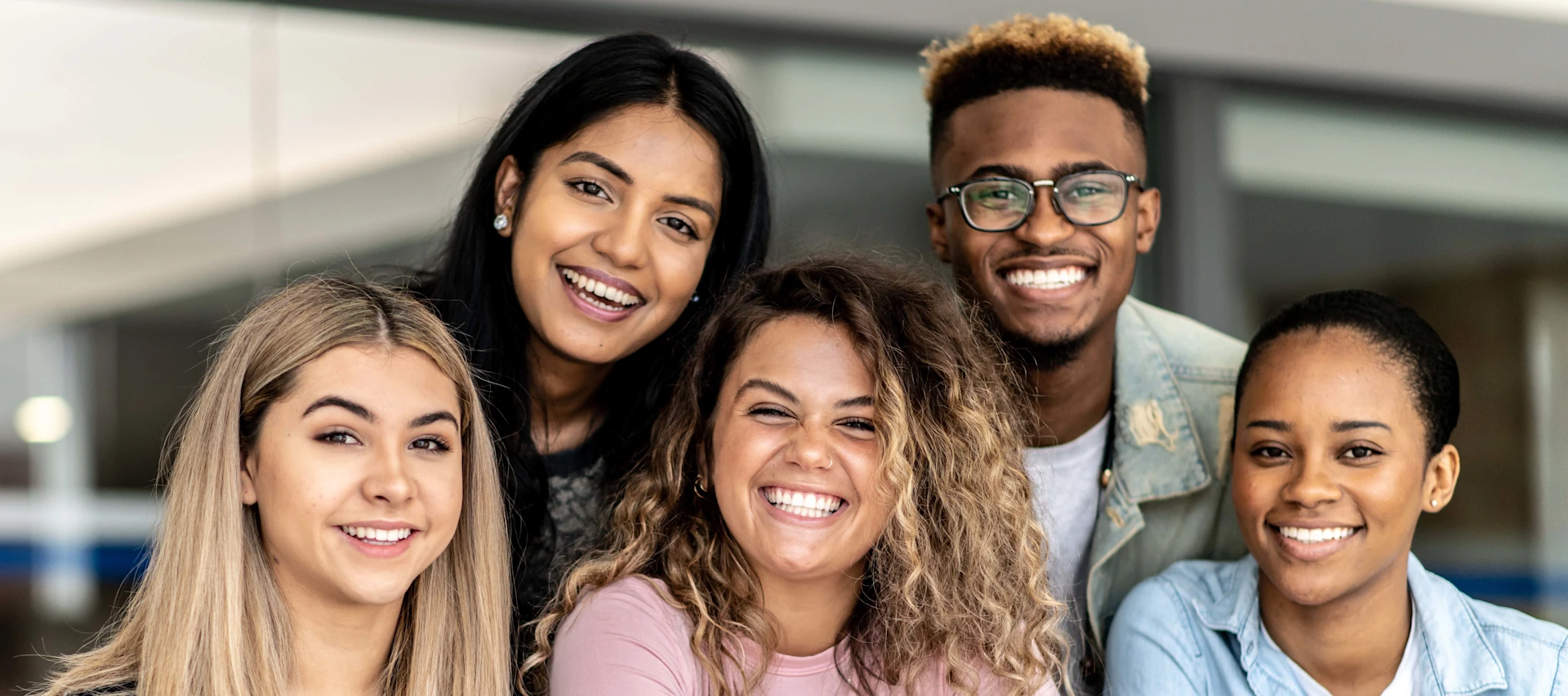 Um grupo de cinco jovens adultos, três mulheres e dois homens, sorrindo e inclinando-se em direção à câmera. Eles transmitem uma sensação de união e camaradagem alegre.