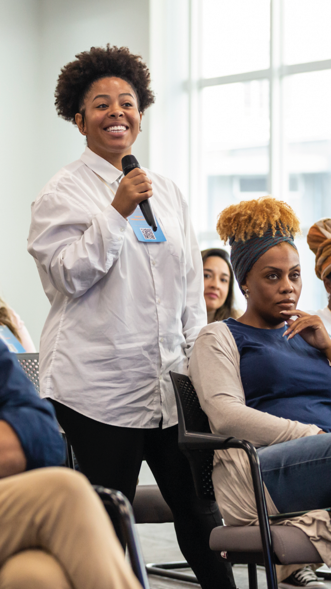Mulher negra sorridente participando de um painel em uma conferência, com outras pessoas ao redor ouvindo atentamente, evento de diversidade e inclusão.