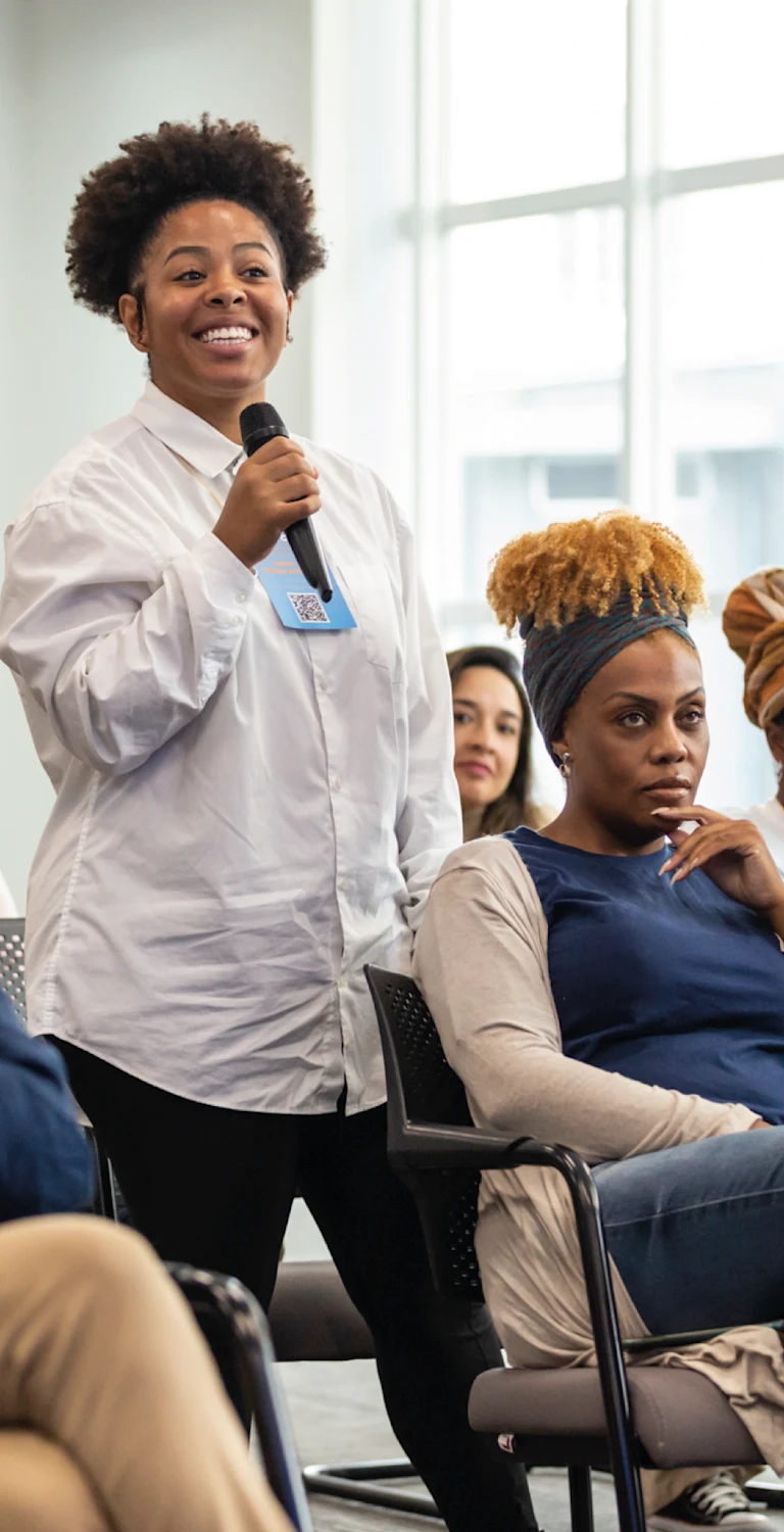 Mulher negra sorridente participando de um painel em uma conferência, com outras pessoas ao redor ouvindo atentamente, evento de diversidade e inclusão.