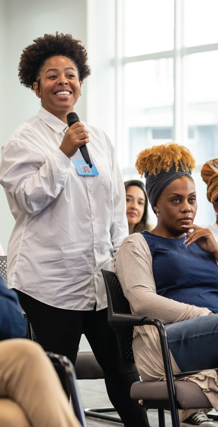Mulher negra sorridente participando de um painel em uma conferência, com outras pessoas ao redor ouvindo atentamente, evento de diversidade e inclusão.