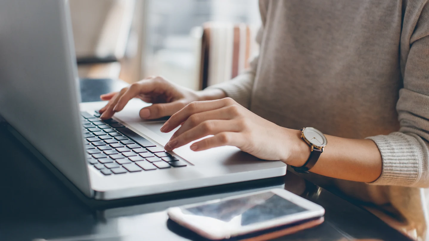 Pessoa digitando em um laptop em uma mesa, com um smartphone próximo. Apenas as mãos e parte do torso são visíveis.