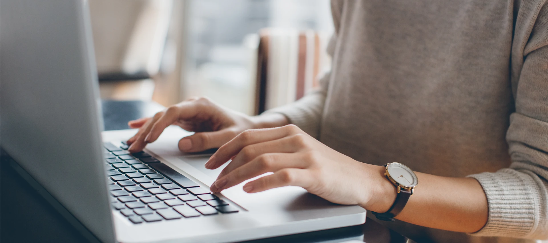 Pessoa digitando em um laptop em uma mesa, com um smartphone próximo. Apenas as mãos e parte do torso são visíveis.