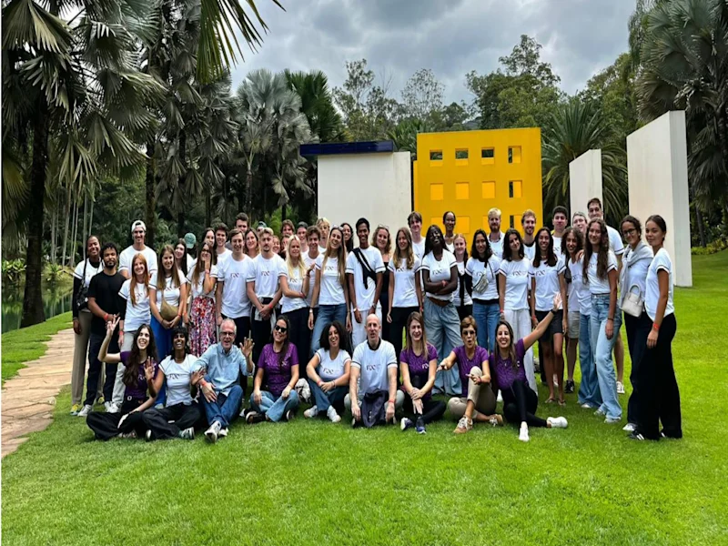 Large group of visitors posing on a lawn at Inhotim Institute, gathered in front of a colorful outdoor artwork, surrounded by tropical gardens and palm trees under a cloudy sky.