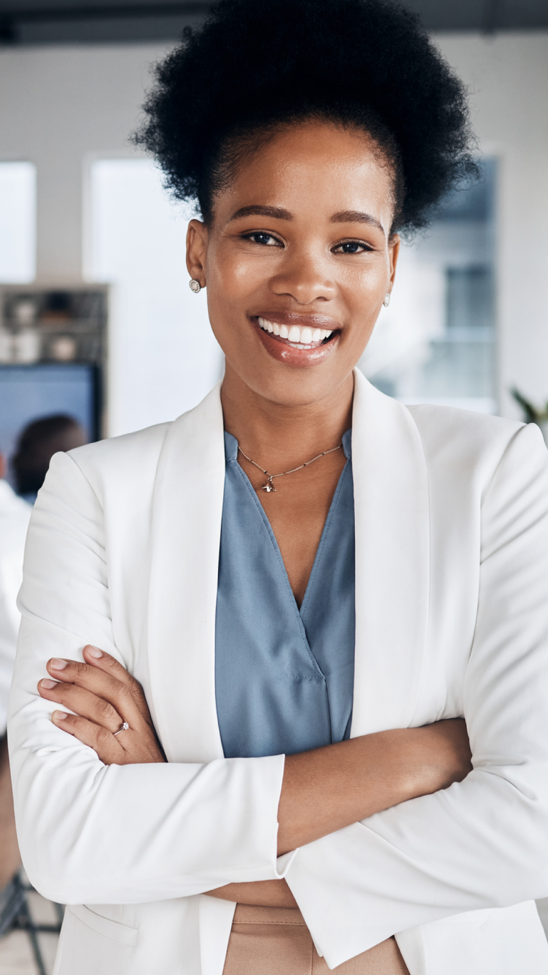 Mulher sorridente de cabelo afro, usando blazer branco e blusa azul, em ambiente de escritório moderno com equipe ao fundo