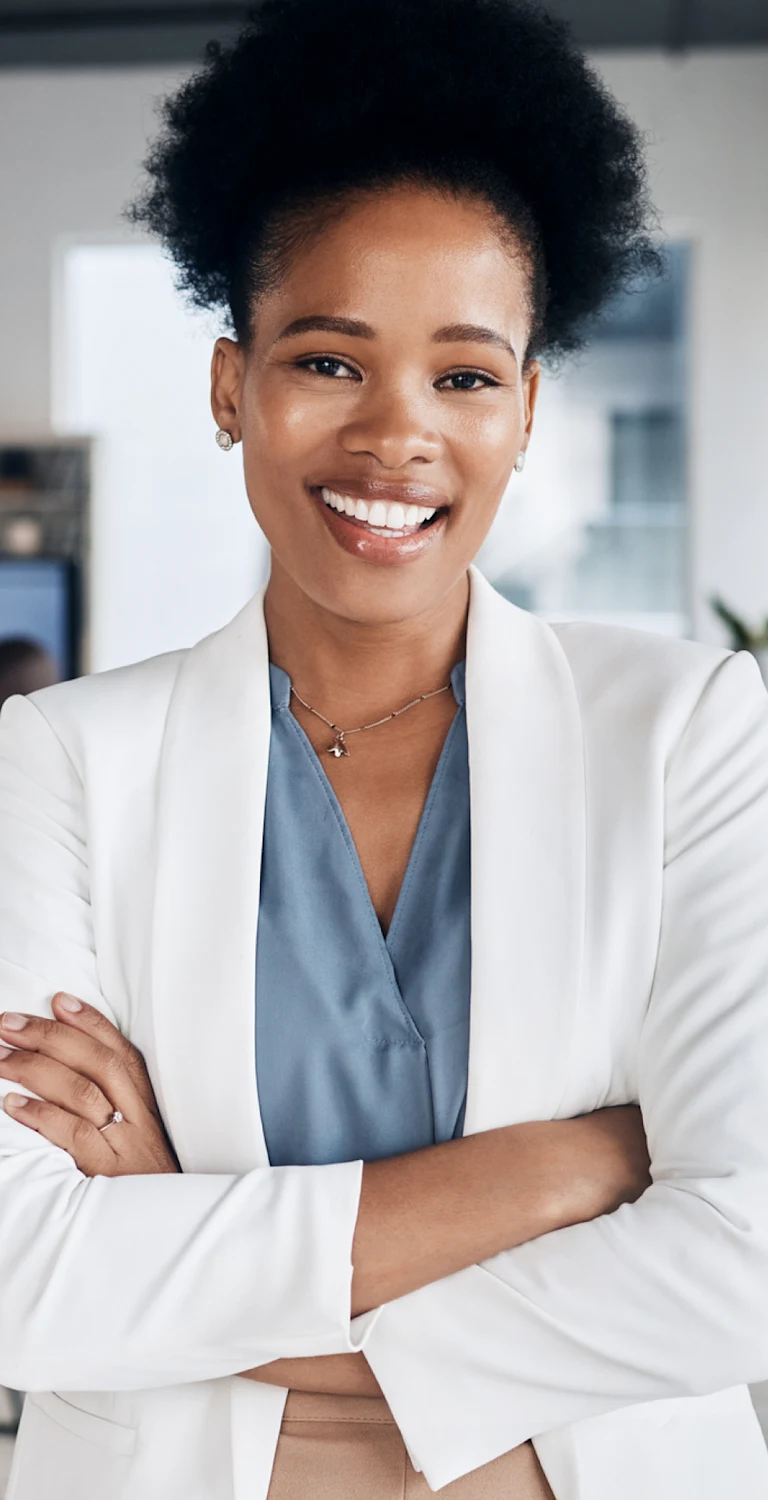 Mulher sorridente de cabelo afro, usando blazer branco e blusa azul, em ambiente de escritório moderno com equipe ao fundo