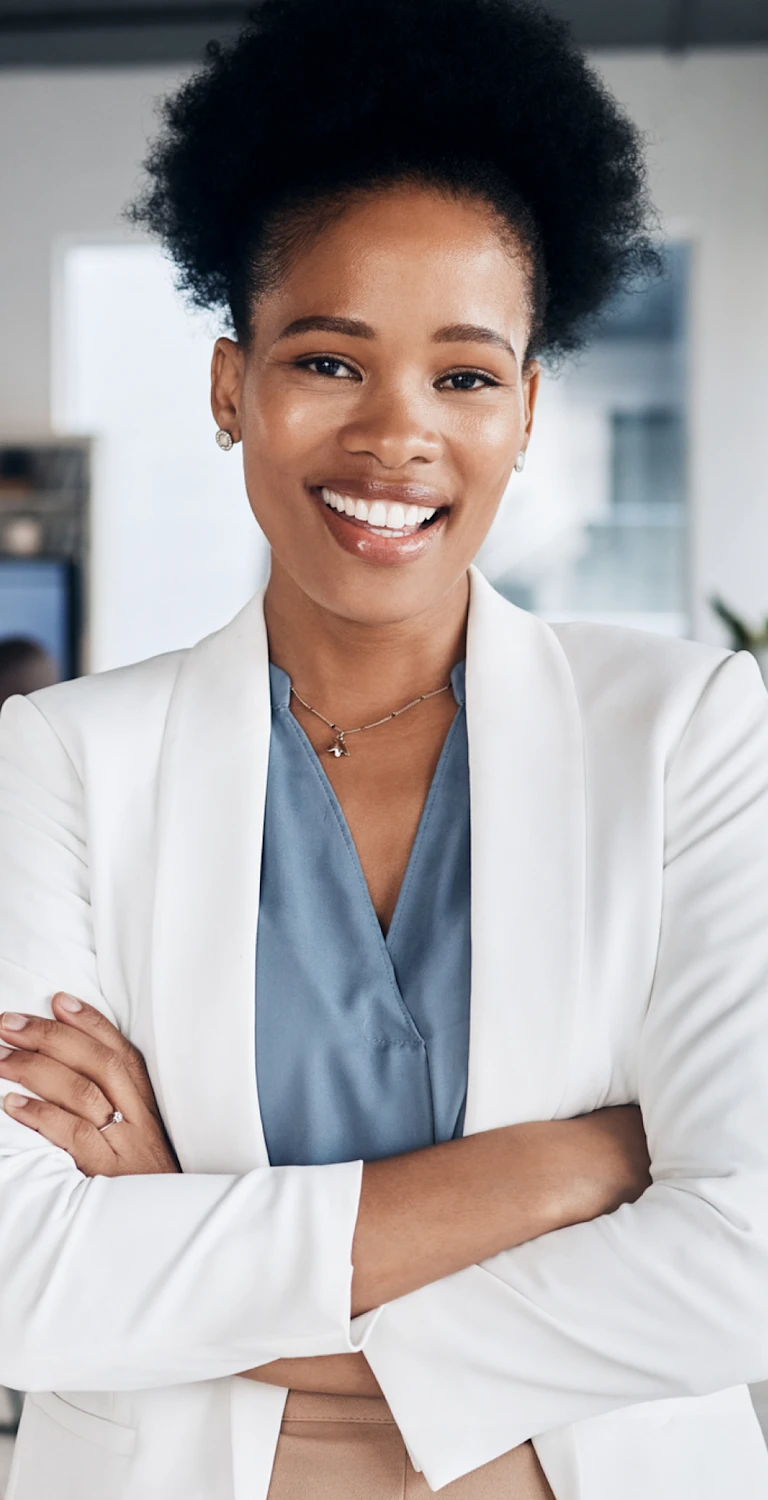 Mulher sorridente de cabelo afro, usando blazer branco e blusa azul, em ambiente de escritório moderno com equipe ao fundo