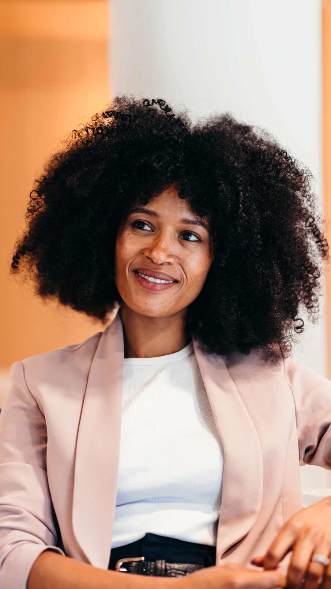 Mulher negra com cabelos cacheados, usando blazer rosa claro e camiseta branca, sorrindo em ambiente de escritório moderno, promovendo empoderamento feminino.