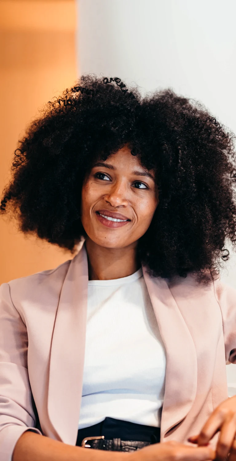 Mulher negra com cabelos cacheados, usando blazer rosa claro e camiseta branca, sorrindo em ambiente de escritório moderno, promovendo empoderamento feminino.