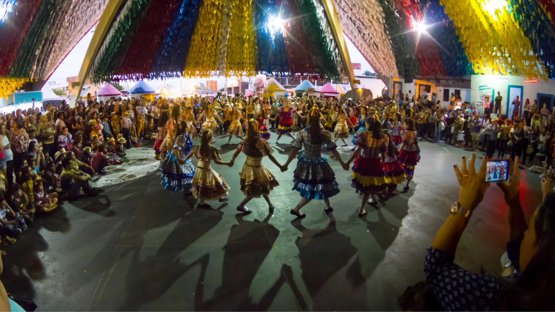 Dancers in colorful traditional costumes perform in a circle at a lively Brazilian Festa Junina celebration, surrounded by a large crowd under a canopy of vibrant hanging decorations.