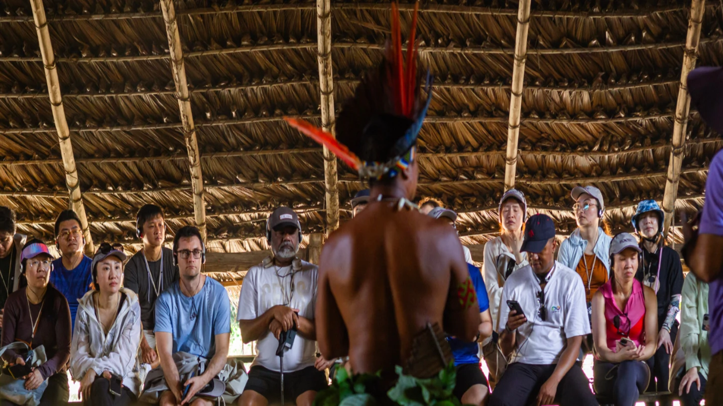 Visitors sit inside a large thatched-roof shelter, watching an Indigenous performer wearing a feathered headdress and leaf skirt. The performer stands with their back to the camera during a cultural demonstration.