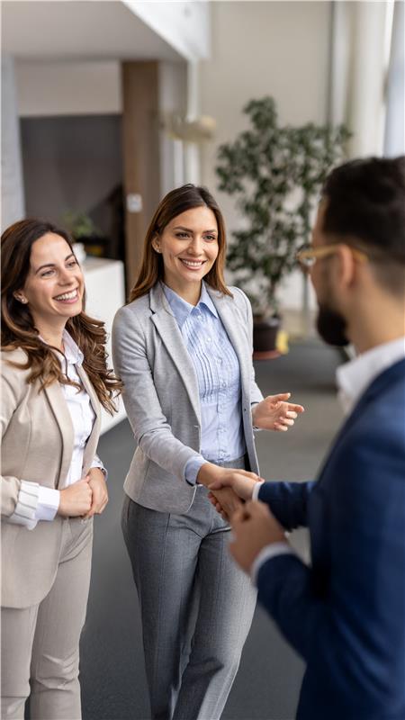 Duas mulheres apertando as mãos em um gesto de parceria, um homem sorri mais ao fundo