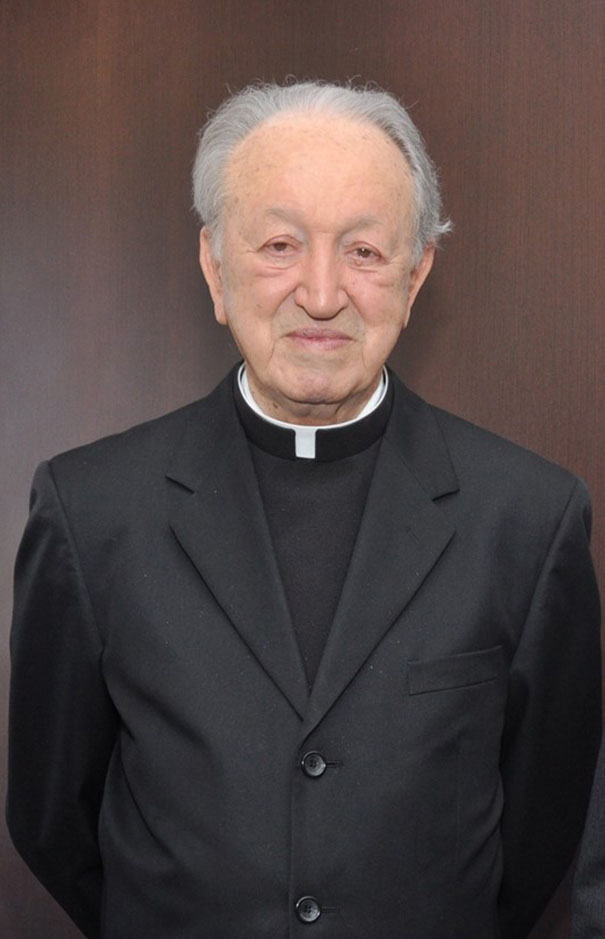 Imagem de um sacerdote idoso, vestido com vestuário clerical preto, com cabelo branco e expressão tranquila, posando em ambiente interno.