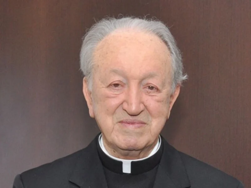 Imagem de um sacerdote idoso, vestido com vestuário clerical preto, com cabelo branco e expressão tranquila, posando em ambiente interno.