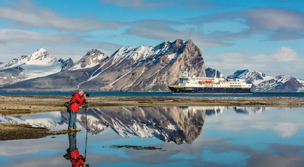 A traveler photographs the landscape in front of mountains and the National Geographic Explorer, Norway