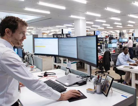 An office worker stands at a desk with multiple monitors, while other employees work in the background of an open-plan office.