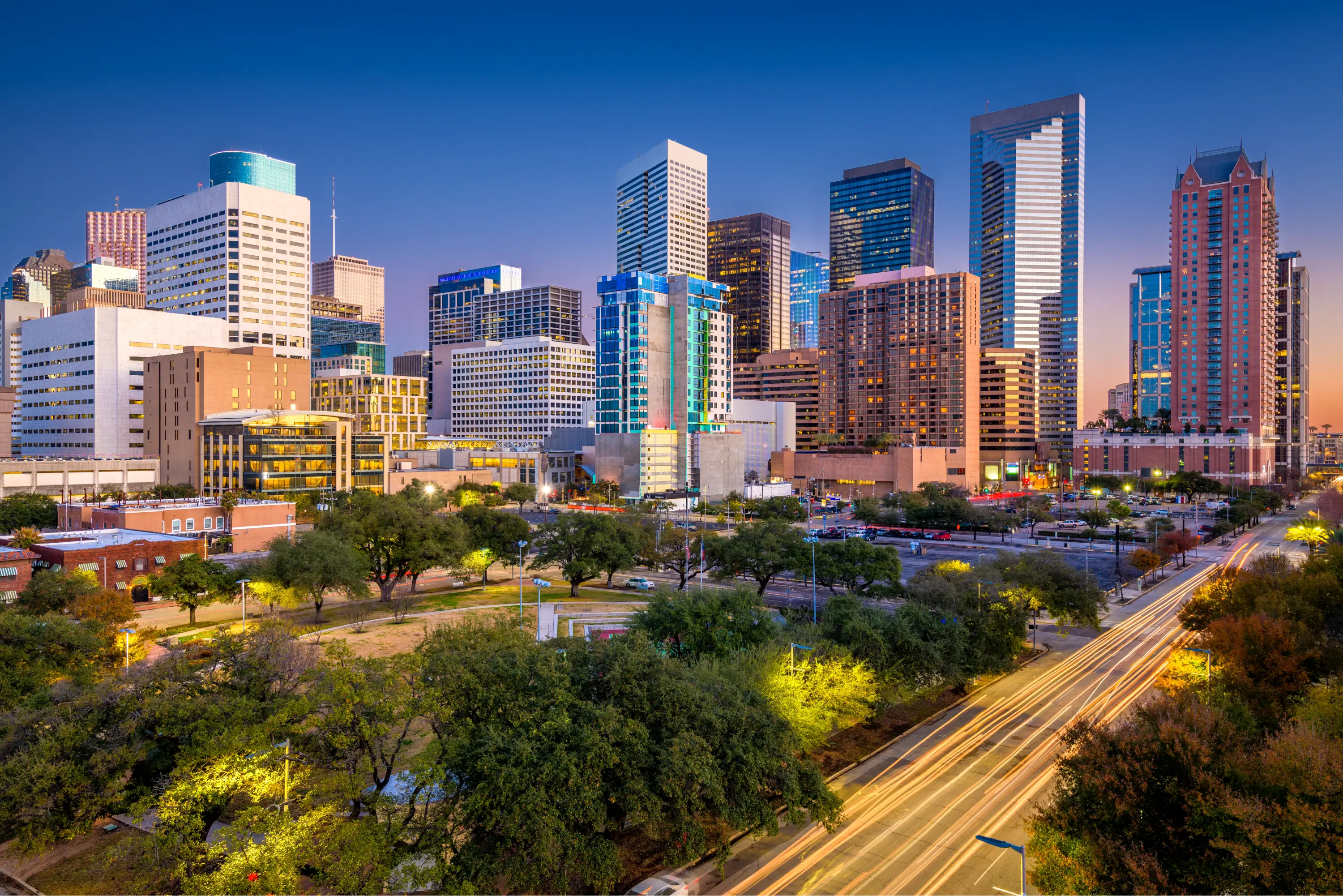 Skyline of downtown Houston, Texas, at dusk with bright lights from cars streaking along the road and modern skyscrapers rising above a mix of green spaces and urban structures.