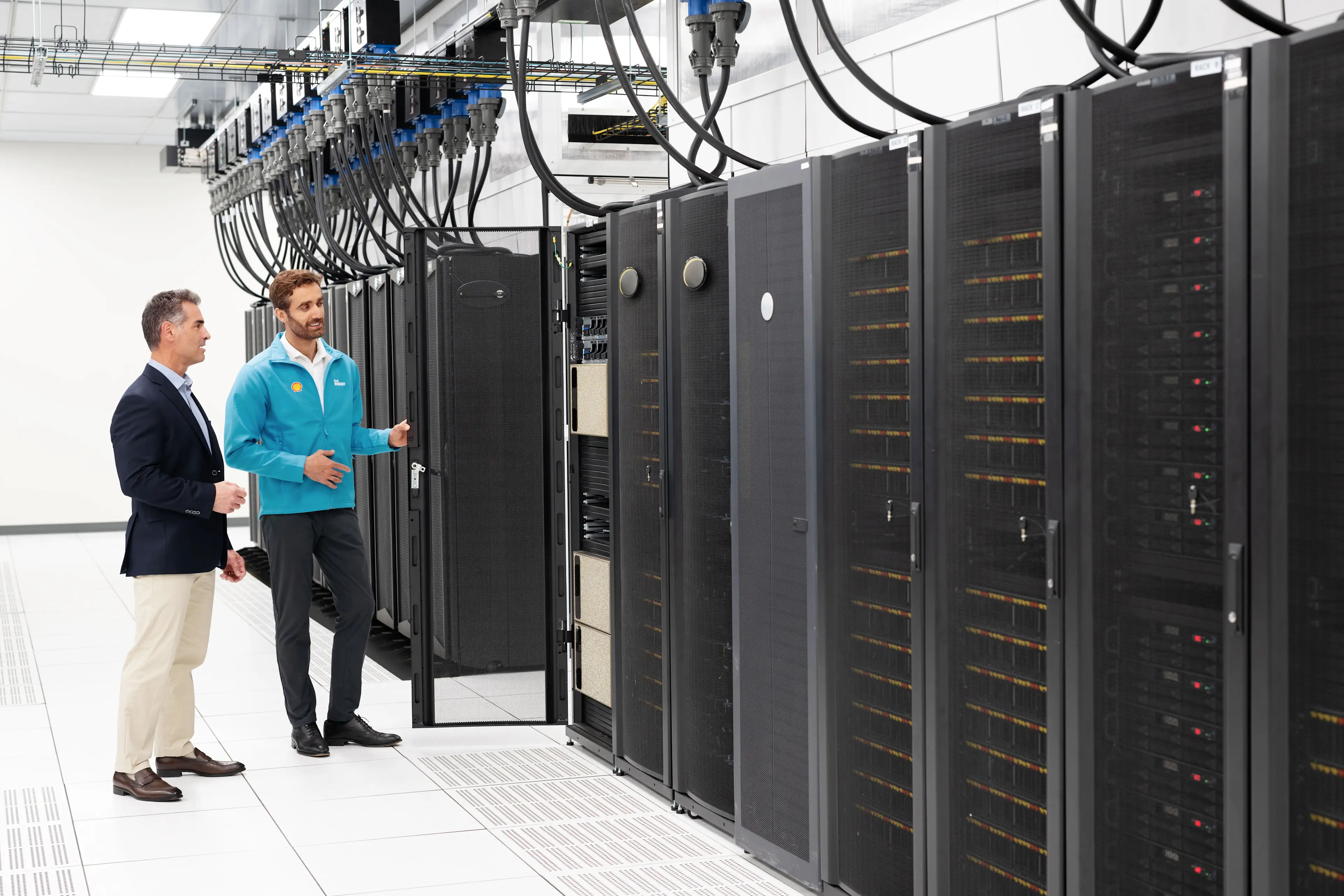 Two men inspecting a row of server racks in a high-tech data center, discussing the equipment while standing in front of black cabinets connected by large cables.