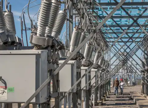 An outdoor electrical substation with rows of high-voltage equipment and two workers walking through the site