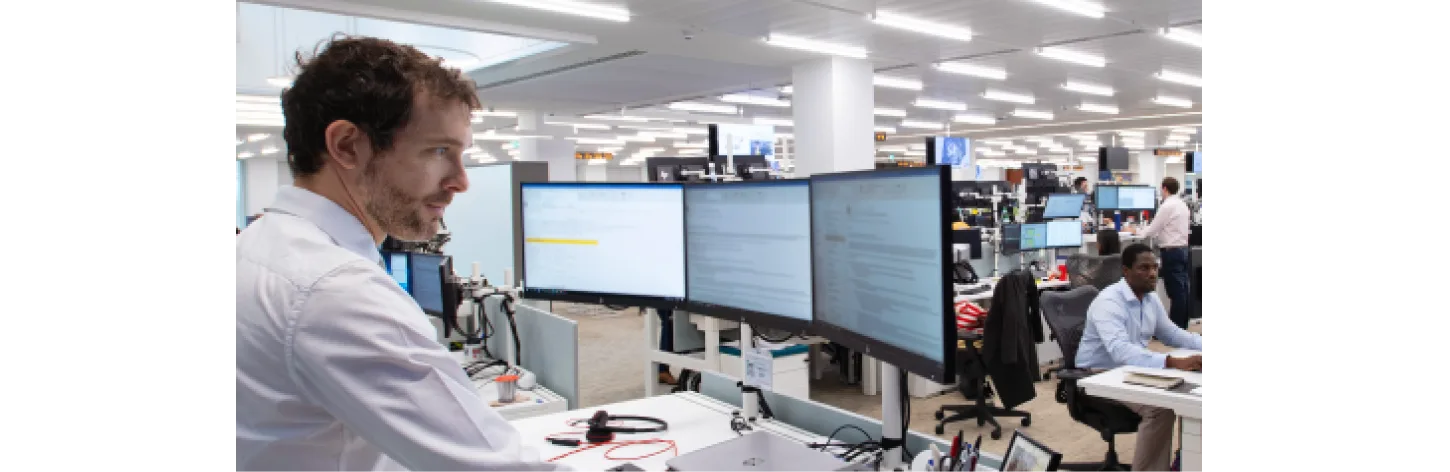 An office worker stands at a desk with multiple monitors, while other employees work in the background of an open-plan office.