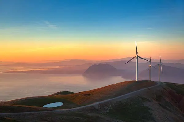 Three wind turbines are positioned along a hilltop at sunset, overlooking a calm body of water and distant mountains.