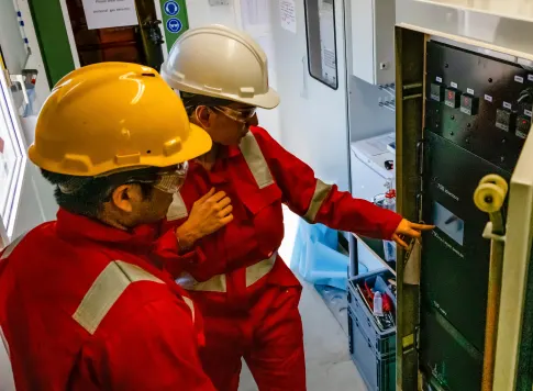 Two workers in safety gear inspecting an electrical control panel inside an industrial facility.