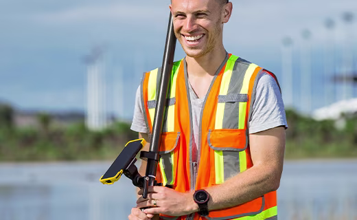 Smiling field user, standing by a pond using data collector and survey pole with hills in background.