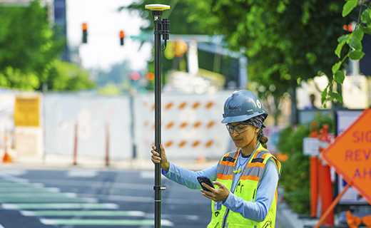 Topógrafa de pie en la calle de una ciudad mirando el smartphone que lleva en la mano mientras sujeta un jalón topográfico con el receptor Trimble DA2 en la parte superior.