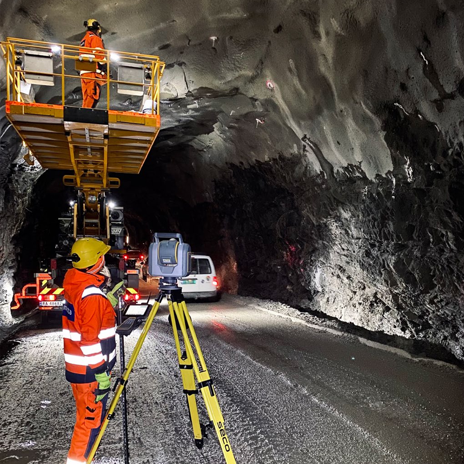 Two workers in high-visibility vests using a Trimble SX12 total station and scanner set up on a tripod