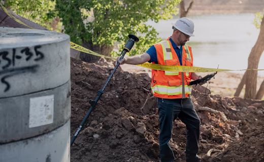 A surveyor in a hard hat and safety vest stands in a dirt trench beside a concrete pipe, holding a tilted ST30 smart target in one hand and a surveyor controller in the other. 