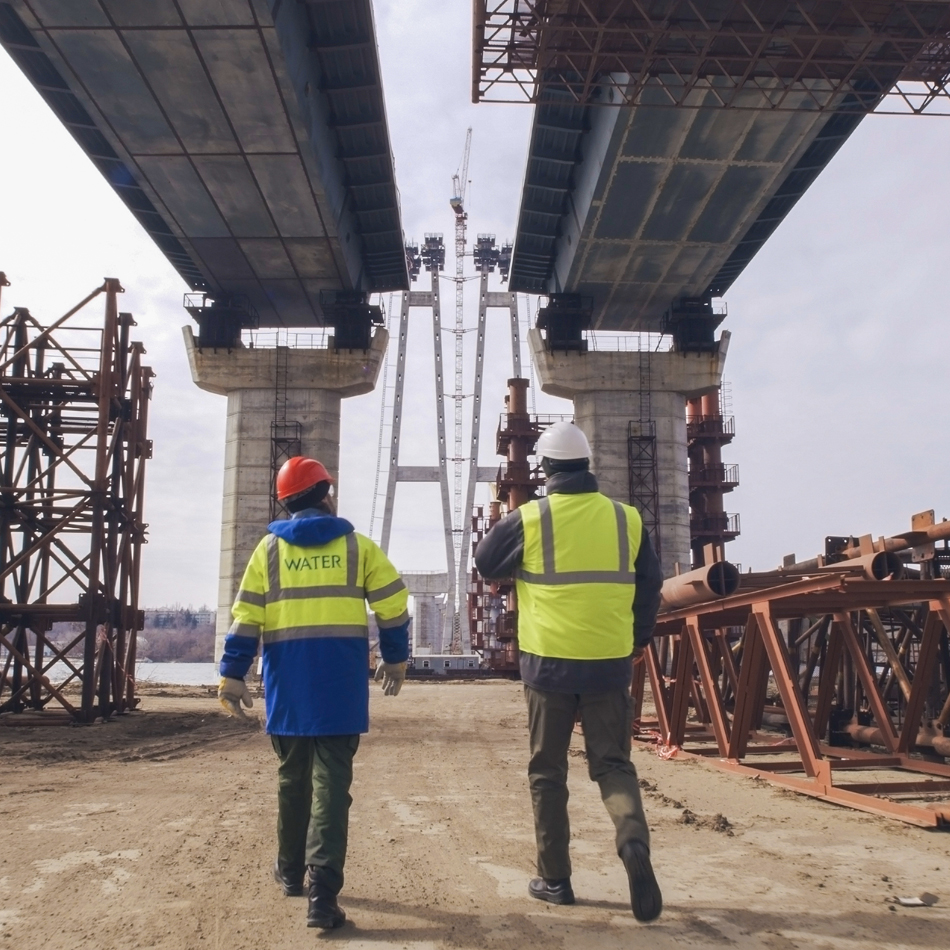 Two construction workers, wearing safety gear, stand beneath a bridge