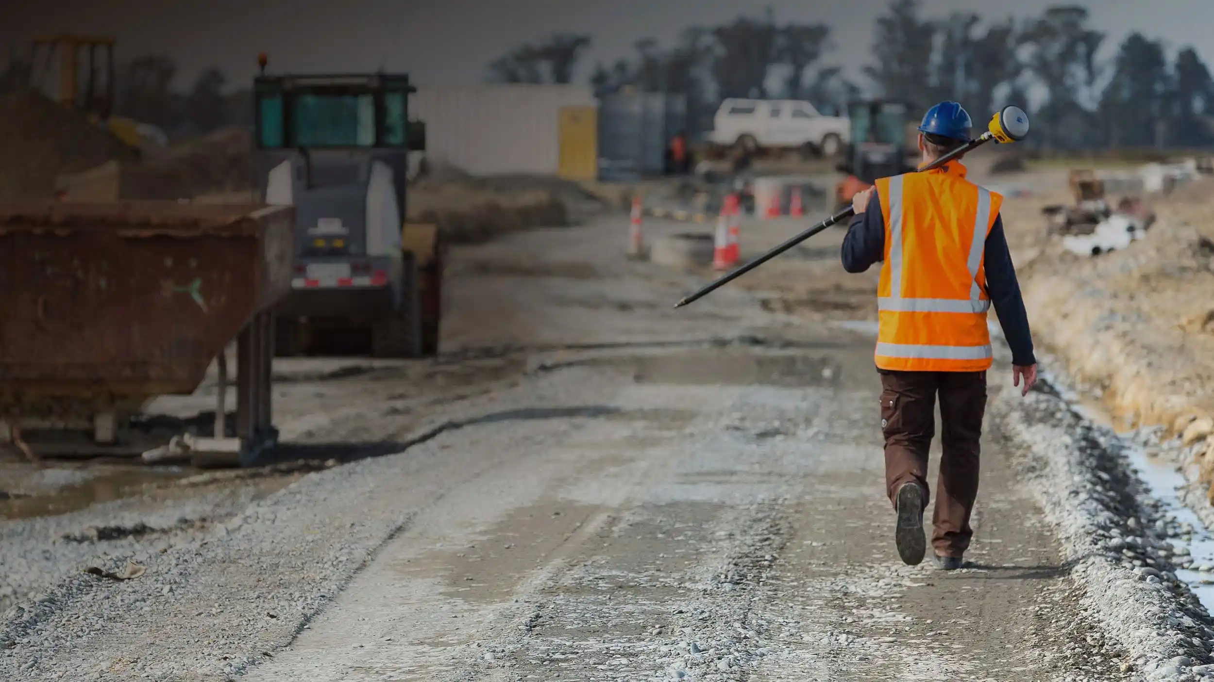 Worker wearing orange safety vest, carrying a Trimble GNSS receiver on a road being repaved.