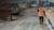 Worker wearing orange safety vest, carrying a Trimble GNSS receiver on a road being repaved.