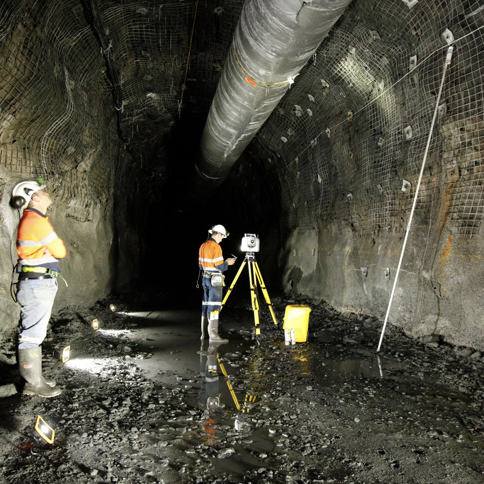 Two workers in high-visibility vests using a Trimble SX12 total station and scanner set up on a tripod